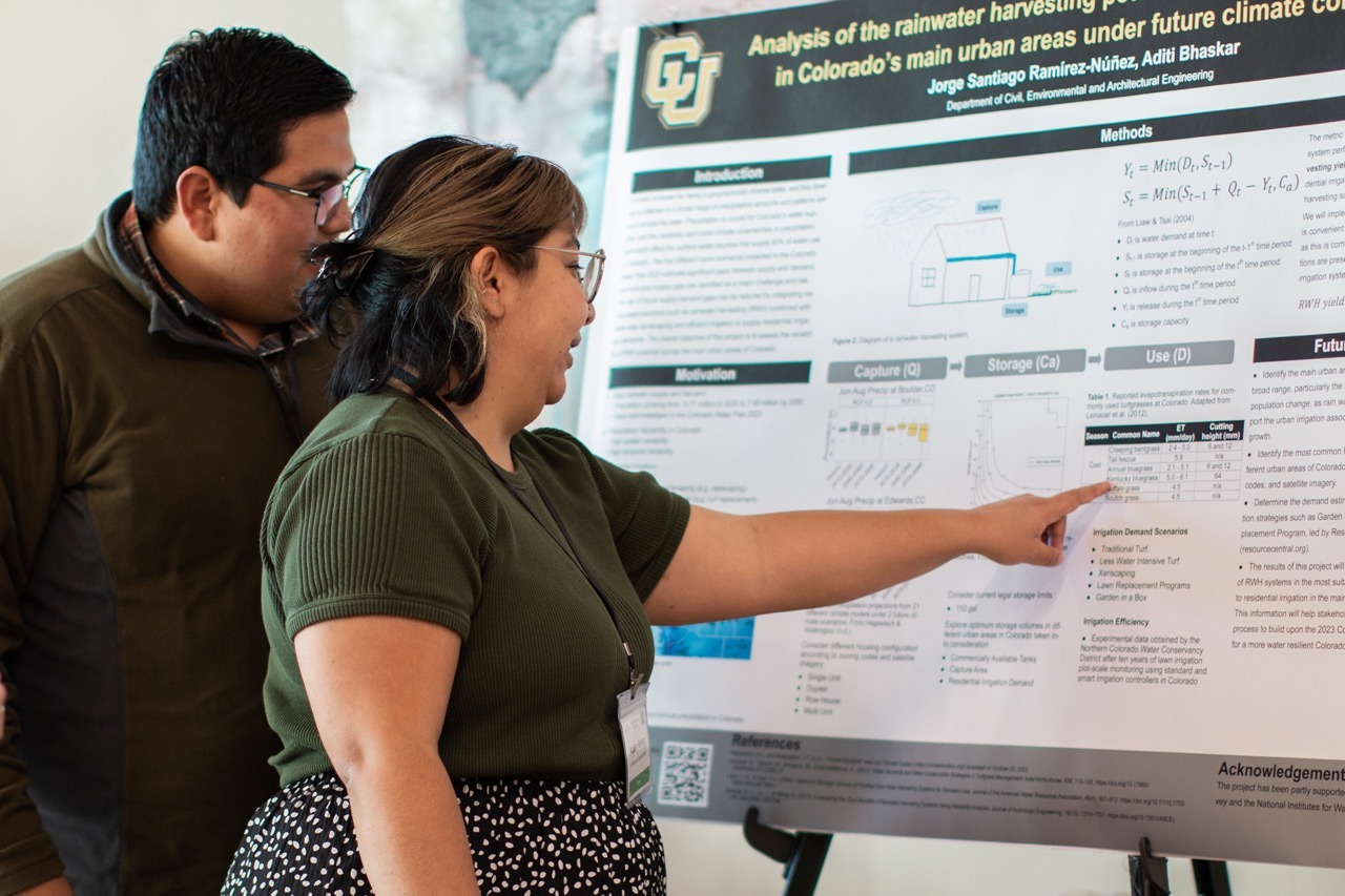 a woman and a man stand next to each other looking at a research poster. The woman points toward some text on the poster.